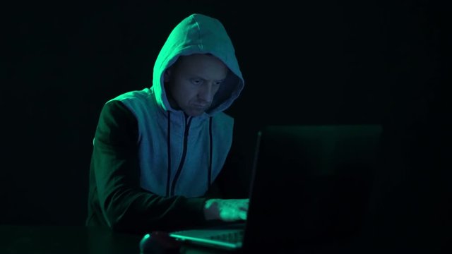 Man a hacker in the hood, at the computer, looks at the monitor and prints codes.