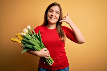 Young blonde woman holding romantic bouquet of tulips flowers over yellow background smiling doing phone gesture with hand and fingers like talking on the telephone. Communicating concepts.