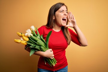 Young blonde woman holding romantic bouquet of tulips flowers over yellow background shouting and screaming loud to side with hand on mouth. Communication concept.