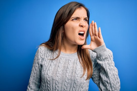 Beautiful young woman wearing casual wool sweater standing over blue isolated background shouting and screaming loud to side with hand on mouth. Communication concept.