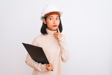 Chinese architect woman wearing helmet holding clipboard over isolated white background serious face thinking about question, very confused idea