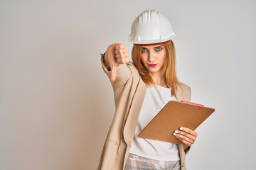 Redhead caucasian business woman wearing safety helmet and holding clipboard with angry face, negative sign showing dislike with thumbs down, rejection concept