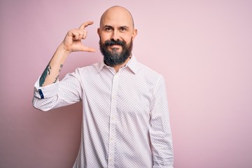 Handsome bald man with beard wearing elegant shirt over isolated pink background smiling and confident gesturing with hand doing small size sign with fingers looking and the camera. Measure concept.