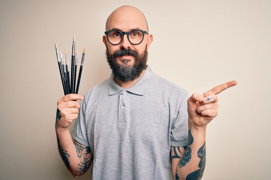 Handsome bald artist man with beard and tattoo painting using painter brushes very happy pointing with hand and finger to the side