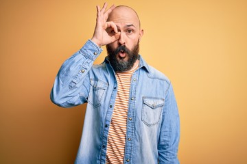 Obraz premium Handsome bald man with beard wearing casual denim jacket and striped t-shirt doing ok gesture shocked with surprised face, eye looking through fingers. Unbelieving expression.