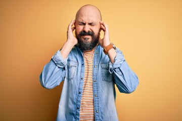 Handsome bald man with beard wearing casual denim jacket and striped t-shirt covering ears with fingers with annoyed expression for the noise of loud music. Deaf concept.