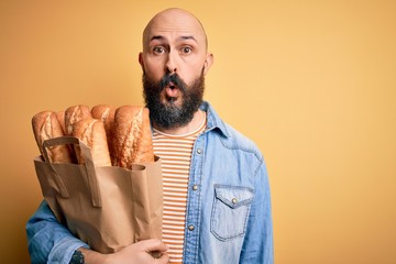 Handsome bald man with beard holding paper bag with bread over yellow background scared in shock...