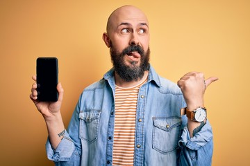 Handsome bald man with beard holding smartphone showing screen over yellow background pointing and showing with thumb up to the side with happy face smiling