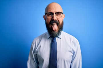 Handsome business bald man with beard wearing elegant tie and glasses over blue background sticking tongue out happy with funny expression. Emotion concept.