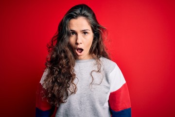 Fototapeta premium Young beautiful woman with curly hair wearing casual sweatshirt over isolated red background afraid and shocked with surprise and amazed expression, fear and excited face.