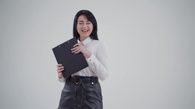 Portrait of a beautiful brunette woman. Attractive lady in white blouse holding black folder and closing her smiling face on the light background. Slow motion.