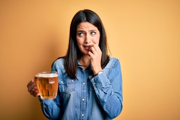 Young woman with blue eyes drinking jar of beer standing over isolated yellow background looking stressed and nervous with hands on mouth biting nails. Anxiety problem.