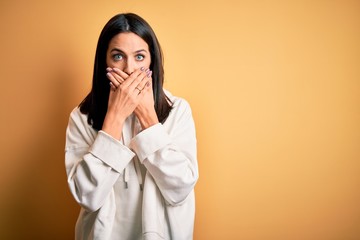 Young brunette sportswoman with blue eyes wearing training sweatshirt over yellow background shocked covering mouth with hands for mistake. Secret concept.