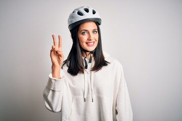 Young cyclist woman with blue eyes wearing bike helmet over isolated white background smiling with happy face winking at the camera doing victory sign. Number two.