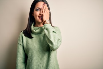 Young brunette woman with blue eyes wearing turtleneck sweater over white background covering one eye with hand, confident smile on face and surprise emotion.