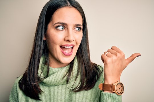 Young brunette woman with blue eyes wearing turtleneck sweater over white background pointing and showing with thumb up to the side with happy face smiling