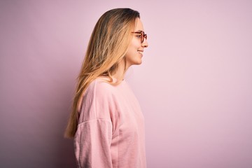 Beautiful blonde woman with blue eyes wearing sweater and glasses over pink background looking to side, relax profile pose with natural face with confident smile.