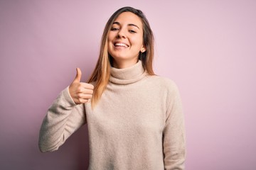 Beautiful blonde woman with blue eyes wearing turtleneck sweater over pink background doing happy thumbs up gesture with hand. Approving expression looking at the camera showing success.