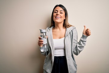 Young beautiful brunette sporty woman drinking bottle of water over isolated white background happy with big smile doing ok sign, thumb up with fingers, excellent sign