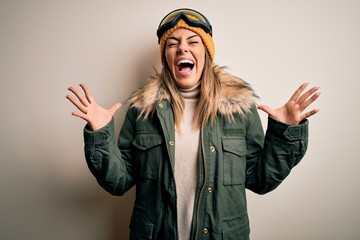 Young brunette skier woman wearing snow clothes and ski goggles over white background celebrating mad and crazy for success with arms raised and closed eyes screaming excited. Winner concept