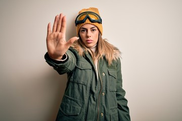 Young brunette skier woman wearing snow clothes and ski goggles over white background doing stop sing with palm of the hand. Warning expression with negative and serious gesture on the face.