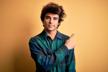 Young handsome man wearing casual shirt standing over isolated yellow background Pointing with hand finger to the side showing advertisement, serious and calm face