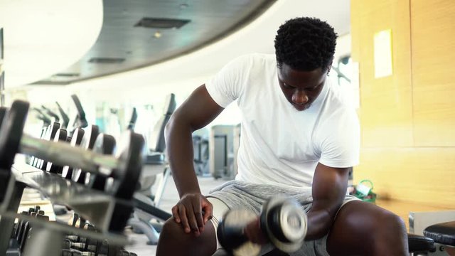 Young African American man sitting and lifting a dumbbell close to the rack at gym. Male weight training person doing a biceps curl in fitness center.
