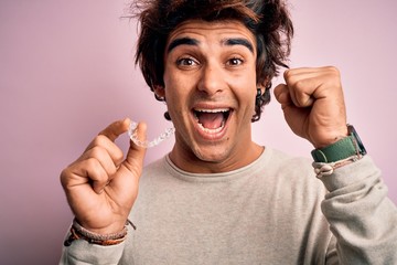 Young handsome man holding aligner standing over isolated pink background screaming proud and...