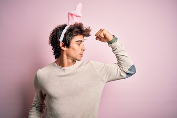 Young handsome man holding easter rabbit ears standing over isolated pink background showing arms muscles smiling proud. Fitness concept.