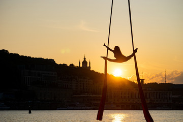 Silhouette of a flexible woman acrobat on aerial silk during a sunset on Kiev city background....