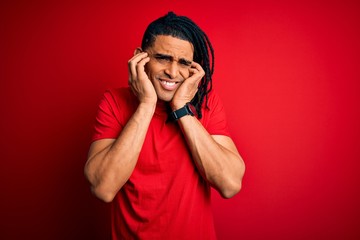 Young handsome african american afro man with dreadlocks wearing red casual t-shirt covering ears with fingers with annoyed expression for the noise of loud music. Deaf concept.