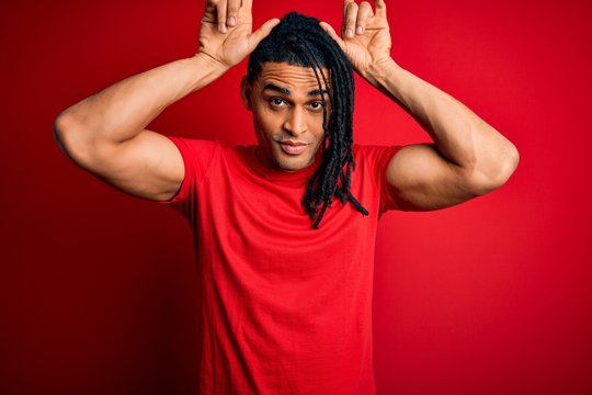 Young handsome african american afro man with dreadlocks wearing red casual t-shirt doing funny gesture with finger over head as bull horns