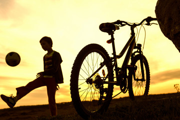 Boy playing with ball in nature, bicycle stands nearby, silhouette of playing child at sunset in countryside
