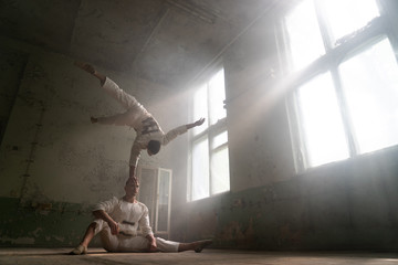 Fototapeta na wymiar Male duo making acrobatic tricks wearing costume of insane people in abandoned room 