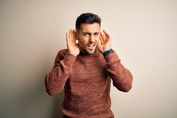 Young handsome man wearing casual sweater standing over isolated white background Trying to hear both hands on ear gesture, curious for gossip. Hearing problem, deaf