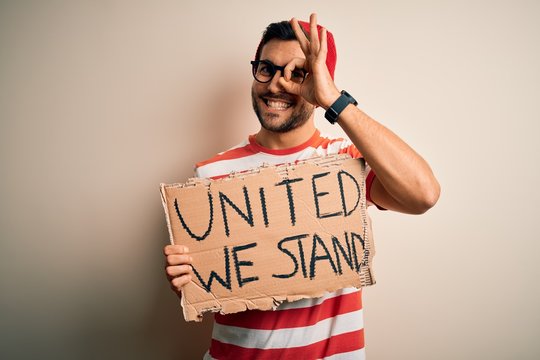 Handsome activist man protesting wearing glasses holding cardboard with unity message with happy face smiling doing ok sign with hand on eye looking through fingers