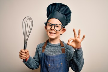 Young little caucasian cook kid wearing chef uniform and hat using manual whisk doing ok sign with...