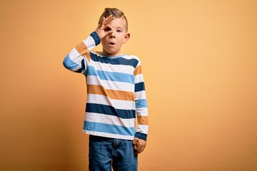 Young little caucasian kid with blue eyes wearing colorful striped shirt over yellow background doing ok gesture shocked with surprised face, eye looking through fingers. Unbelieving expression.