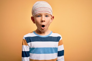 Young little caucasian kid injured wearing medical bandage on head over yellow background afraid...