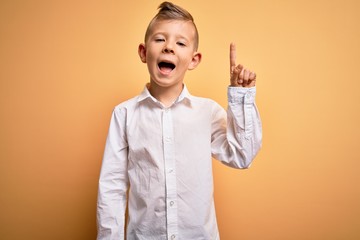 Young little caucasian kid with blue eyes wearing elegant white shirt over yellow background pointing finger up with successful idea. Exited and happy. Number one.