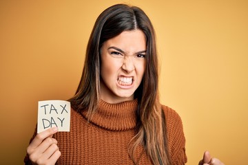 Young beautiful girl holding reminder paper with tax day message over yellow background annoyed and frustrated shouting with anger, crazy and yelling with raised hand, anger concept