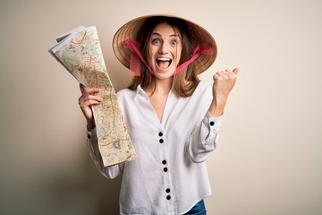Young beautiful redhead woman wearing asian traditional hat holding city map screaming proud and celebrating victory and success very excited, cheering emotion