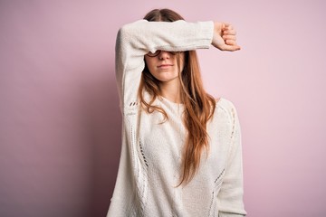 Young beautiful redhead woman wearing casual sweater and glasses over pink background covering eyes with arm, looking serious and sad. Sightless, hiding and rejection concept