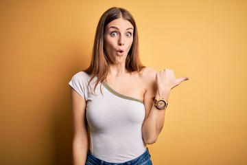 Young beautiful redhead woman wearing casual t-shirt over isolated yellow background Surprised pointing with hand finger to the side, open mouth amazed expression.