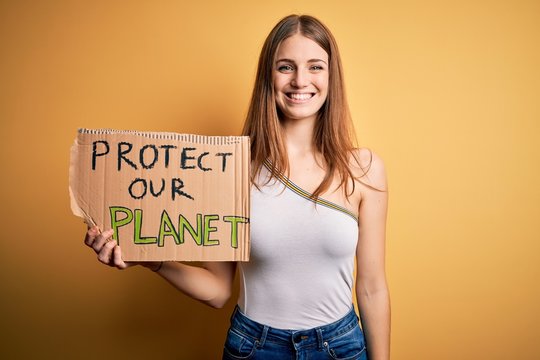 Young redhead woman asking for enviroment holding banner with protect planet message with a happy face standing and smiling with a confident smile showing teeth