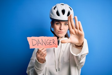 Young beautiful redhead cyclist woman wearing bike helmet holding danger paper message with open hand doing stop sign with serious and confident expression, defense gesture