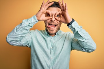 Young business man with blue eyes wearing elegant green shirt over yellow background doing ok gesture like binoculars sticking tongue out, eyes looking through fingers. Crazy expression.