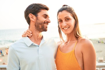 Young beautiful couple on vacation smiling happy and confident. Standing with smile on face hugging at the beach