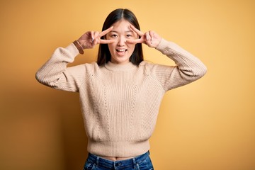 Young beautiful asian woman wearing casual sweater over yellow isolated background Doing peace symbol with fingers over face, smiling cheerful showing victory