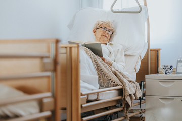 Sick senior woman lies in a hospital bed and reads a book